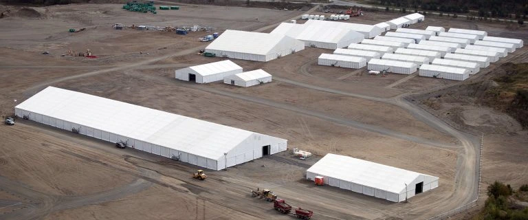 Aerial view of a large-scale temporary base camp on a remote site featuring large industrial warehouse tents and rows of modular accommodation units.