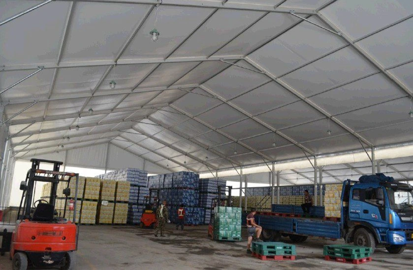 Interior of a busy industrial warehouse tent featuring forklifts loading goods onto a blue truck parked inside the structure.