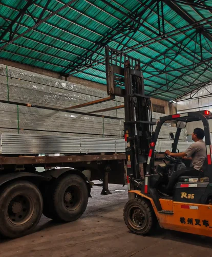 Forklift loading wrapped aluminum tent profiles onto a truck inside a manufacturing facility.