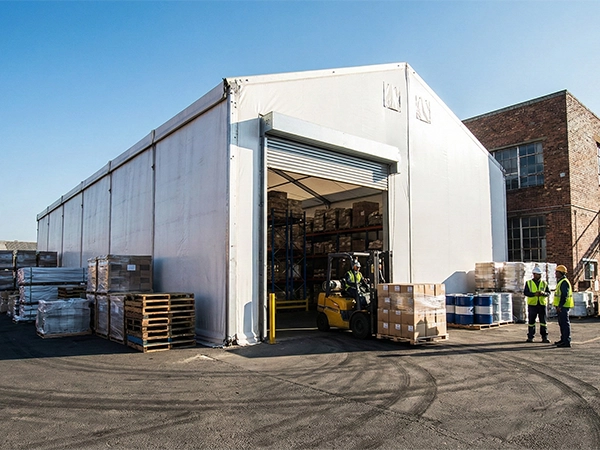 Clear span industrial warehouse tent next to a brick building showing active forklift operations.