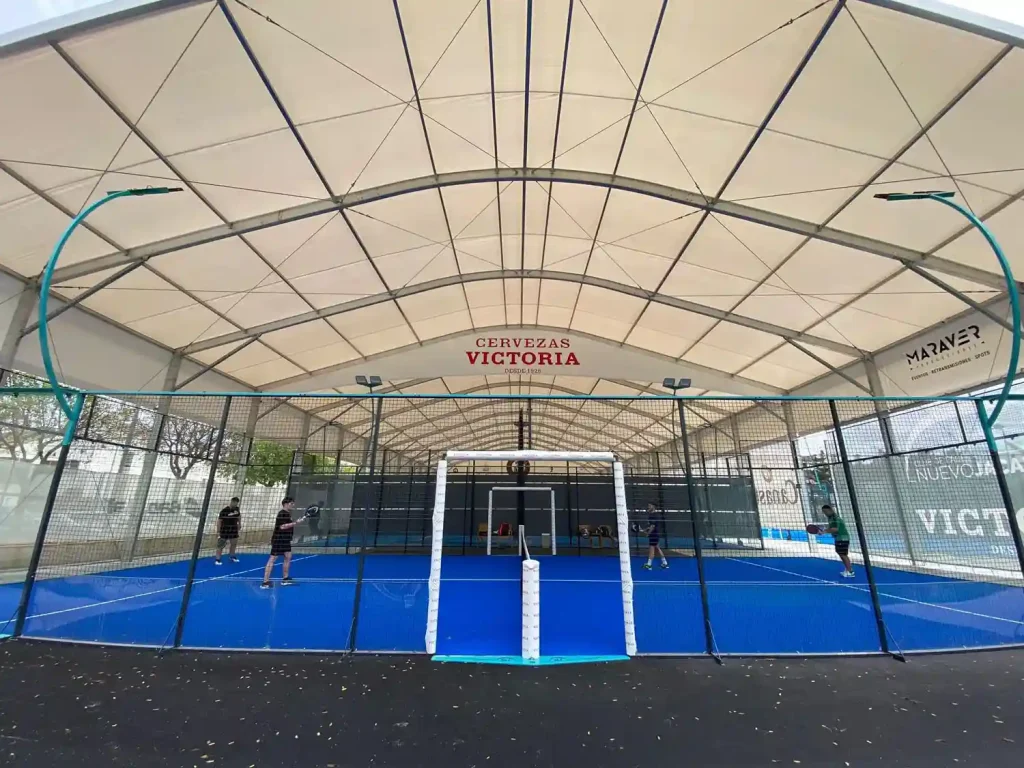 Players competing on a blue padel court inside a large clear span sports tent with a curved roof.