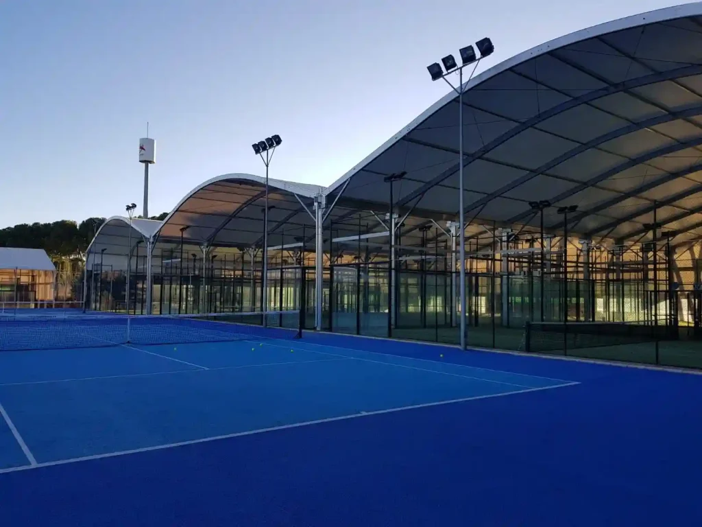 Series of connected curved roof modular sports tents covering outdoor padel courts at dusk.