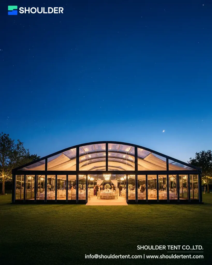 Night view of an illuminated curved roof event tent with glass walls and black aluminum frame.