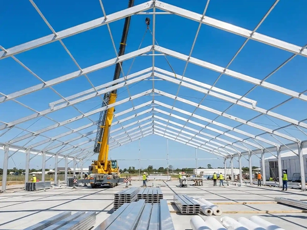 Crane assembling a large aluminum clear span tent frame with PVC rolls in the foreground.