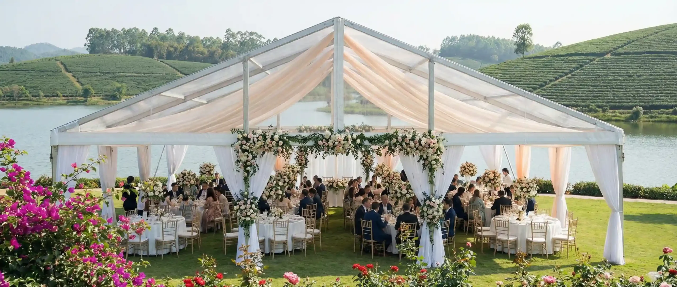 Transparent clear span tent with floral arches and draped lining set up on a lakeside lawn.