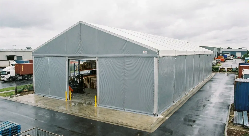 Forklift moving wooden pallets inside a grey clear span storage tent on a wet industrial lot.