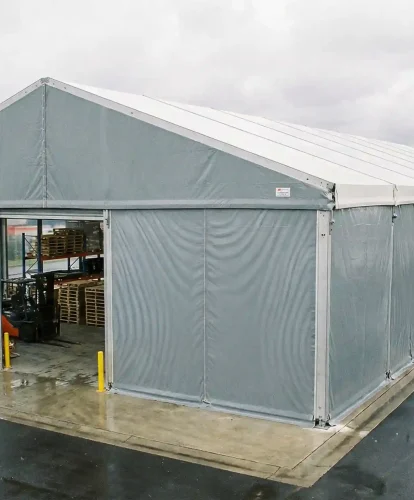 Forklift moving wooden pallets inside a grey clear span storage tent on a wet industrial lot.