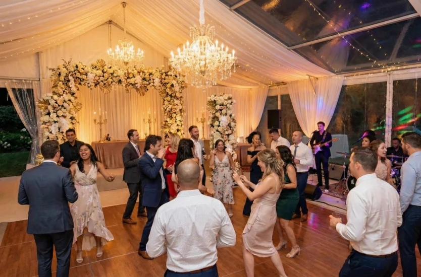 Guests dancing on a wooden floor inside an illuminated clear roof event tent at night.