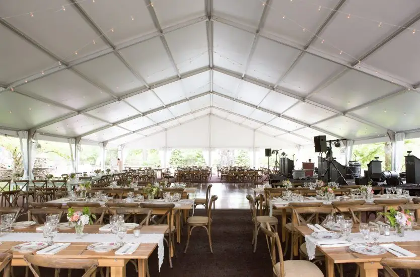Interior of an aluminum clear span tent arranged for a wedding banquet with wooden furniture.