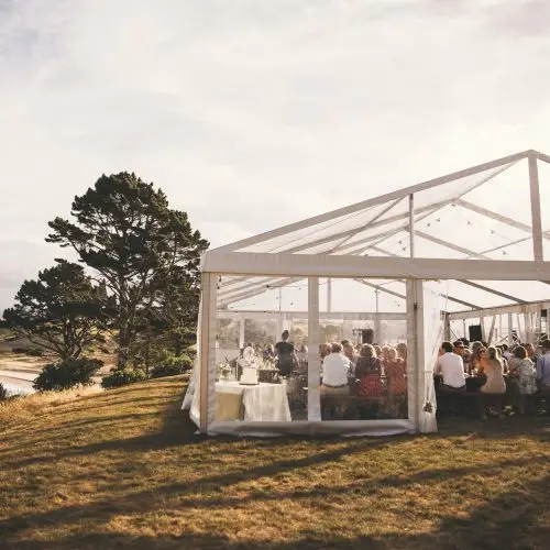 People dining inside a transparent clear span tent situated on a grassy hill at sunset.