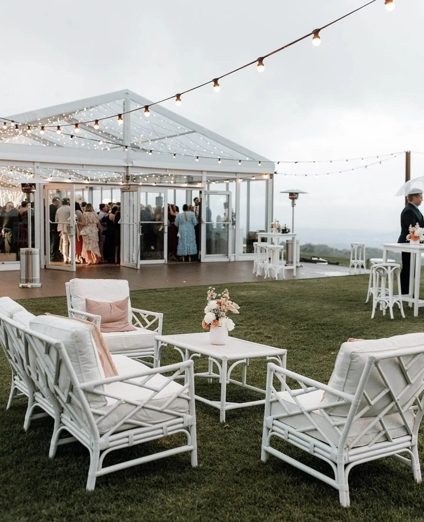 Guests mingling inside a clear span glass event tent with outdoor patio furniture.