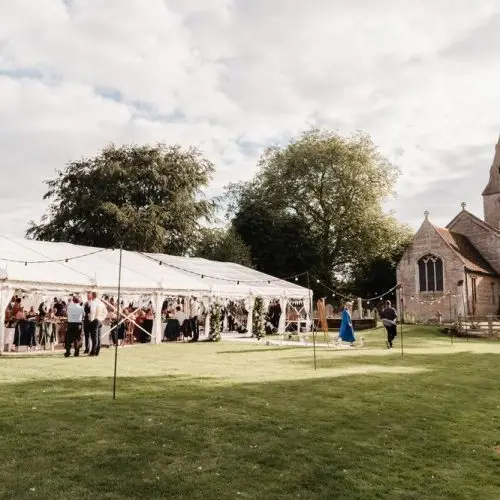 White clear span wedding tent erected on a green lawn beside a historic stone church.