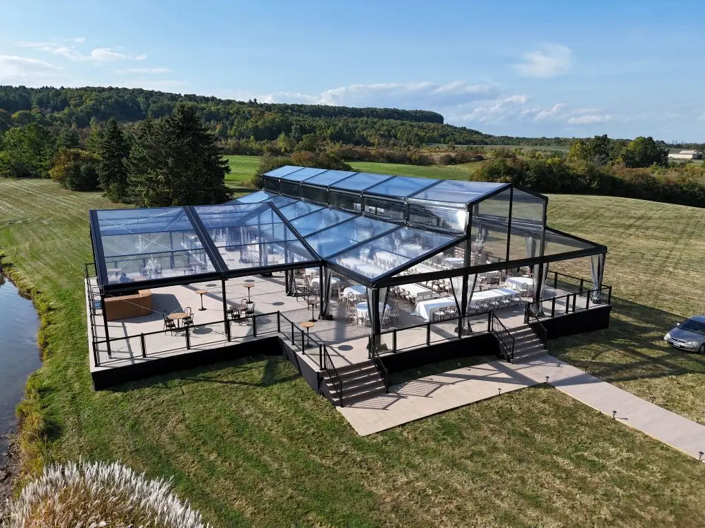 Clear roof event tent with black aluminum frame on a raised deck by a lake.