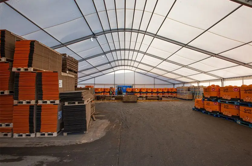 Large curved roof aluminum warehouse tent interior storing stacked pallets and industrial goods.