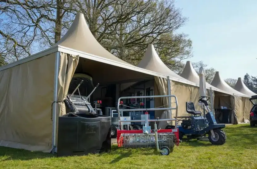 Row of connected tan exhibition tents sheltering agricultural equipment outdoors.