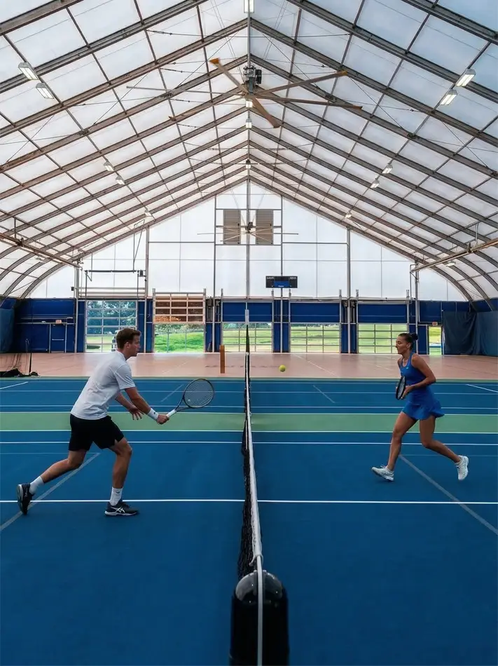 Two people playing tennis inside a clear span sports structure with translucent roof.