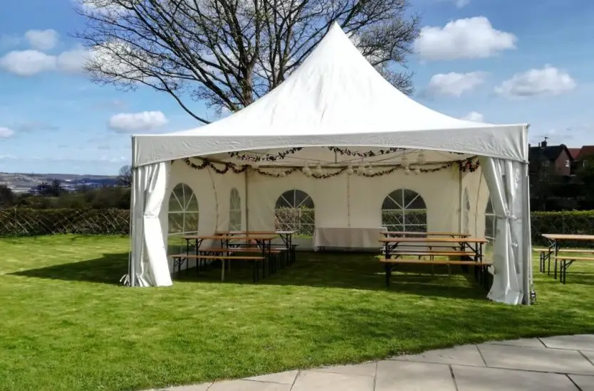 White aluminum frame pagoda tent on grass with clear windows and wooden tables.