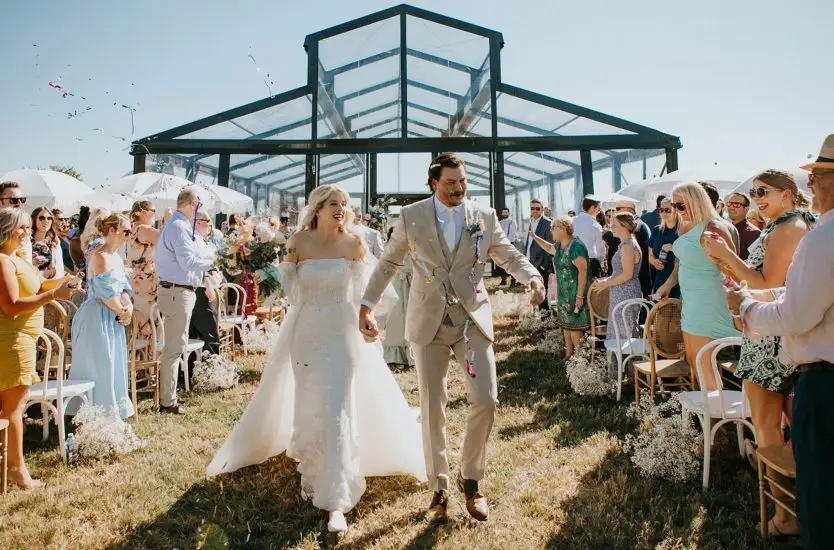 Newlyweds walking out of a black aluminum clear span wedding tent with transparent walls.