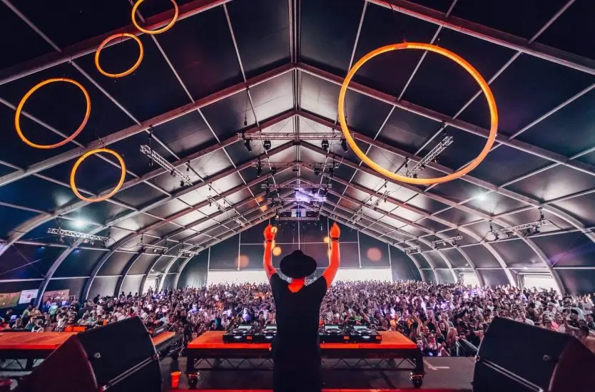 Crowded music festival inside a large aluminum clear span tent with hanging stage lights.